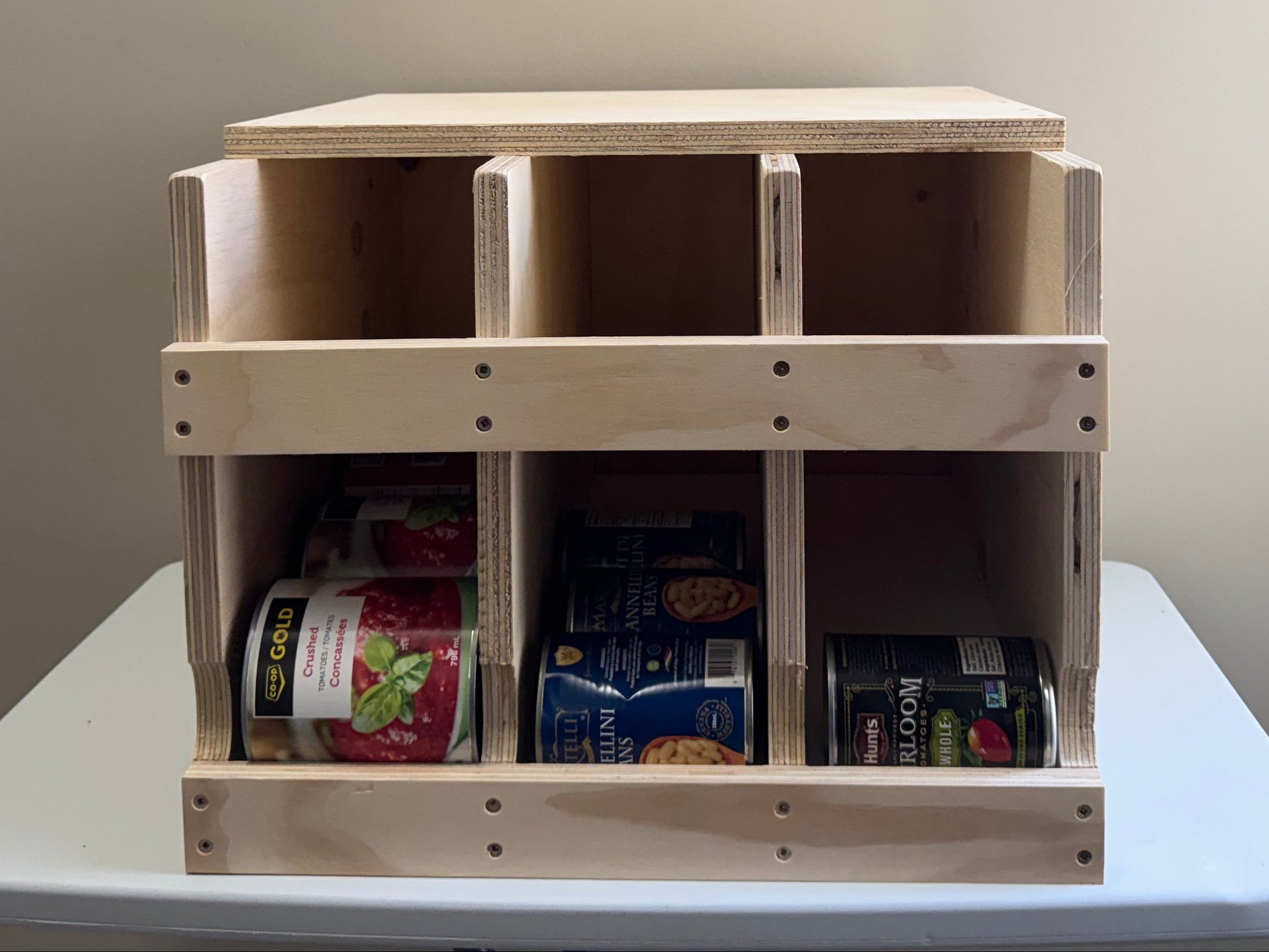 A light natural wood 3-compartment organizer with a lid, shown on a white table against a beige wall. The bottom compartments contain packaged snacks and items.