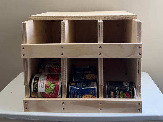 A light natural wood 3-compartment organizer with a lid, shown on a white table against a beige wall. The bottom compartments contain packaged snacks and items.