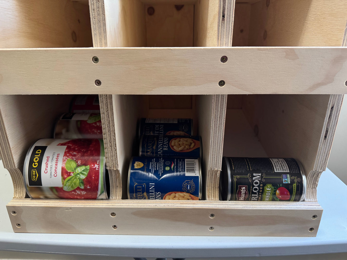 A light natural wood organizer shelf with multiple compartments holding various canned goods, including a can of SP Gold Cherry Pie and a can of Hill's Sweet Home Canned Soup.