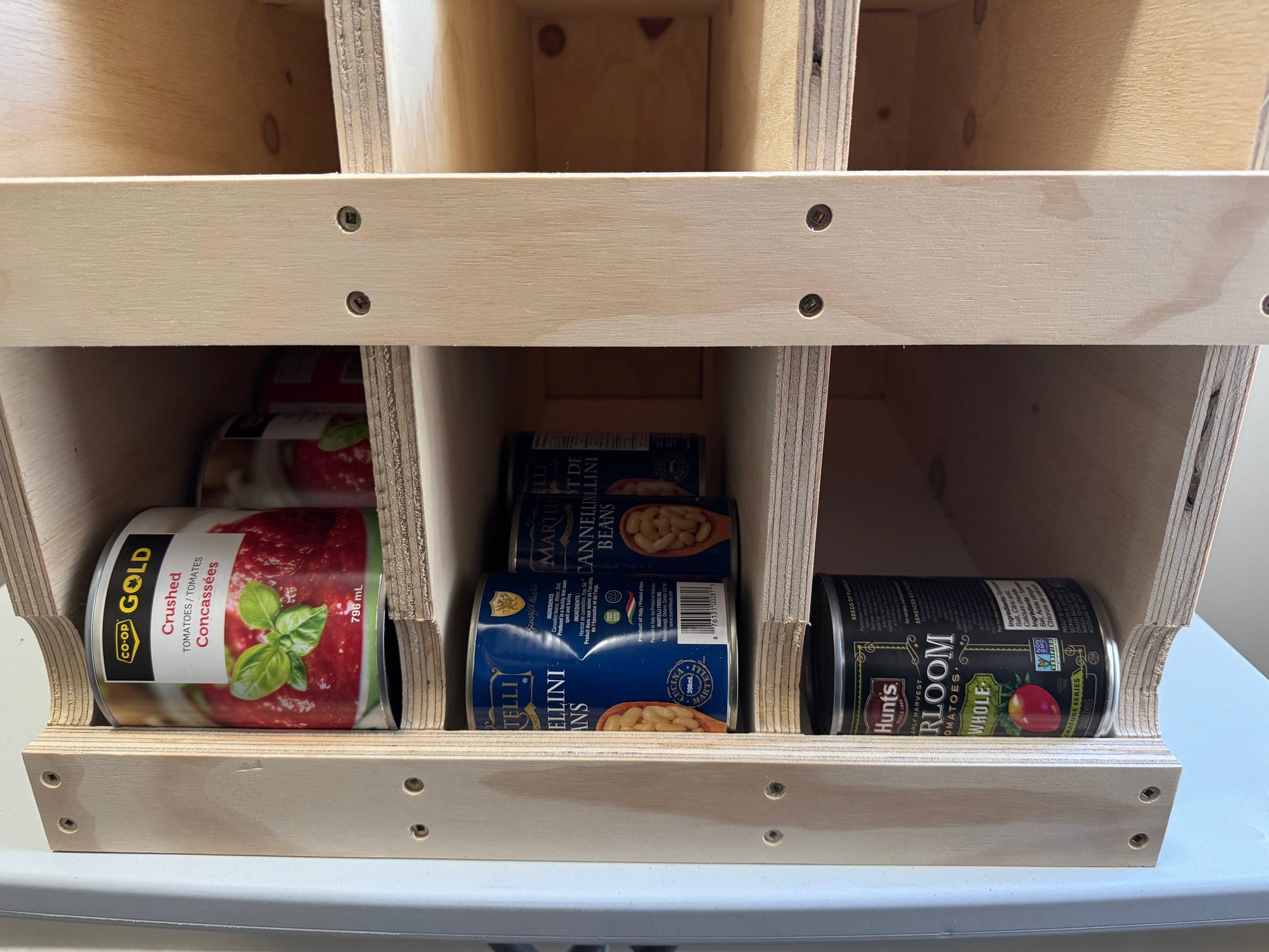 A light natural wood organizer shelf with multiple compartments holding various canned goods, including a can of SP Gold Cherry Pie and a can of Hill's Sweet Home Canned Soup.