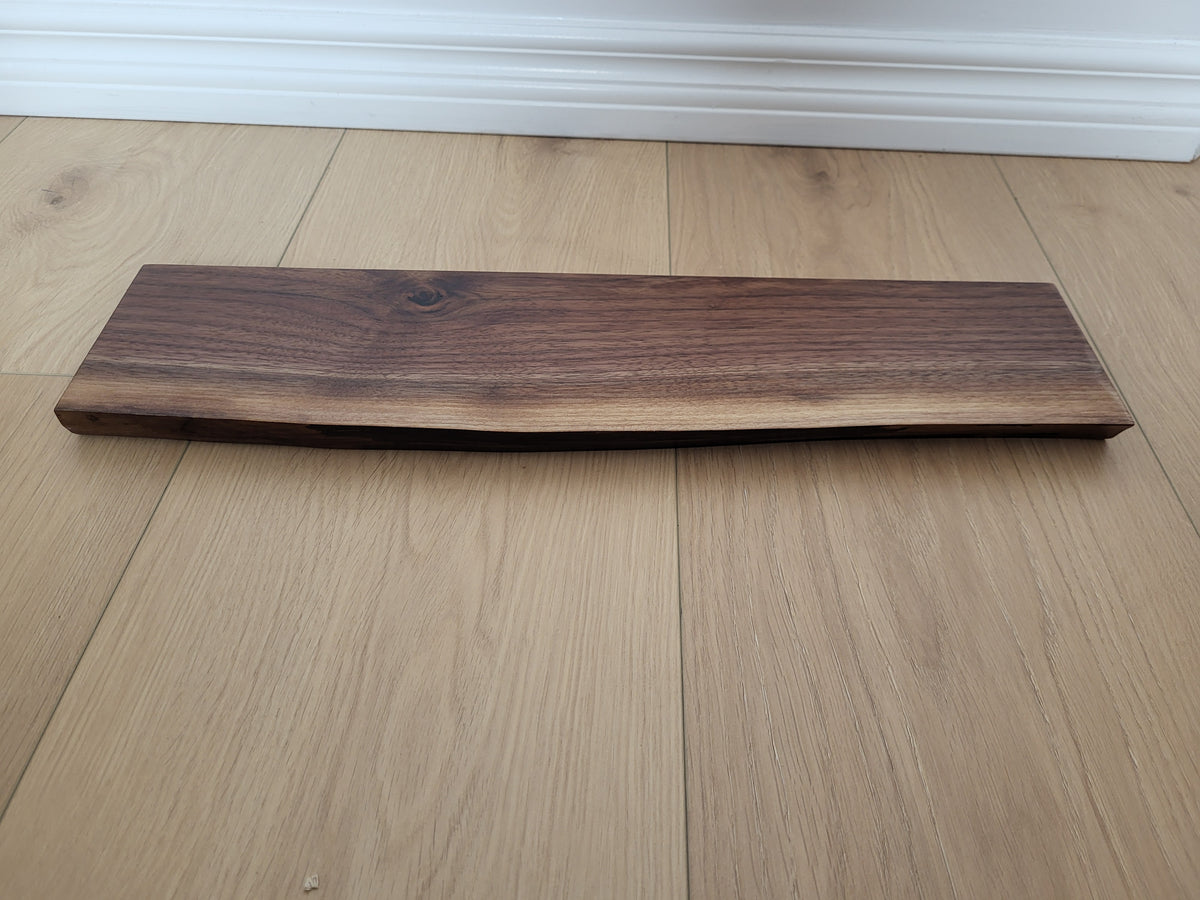A rectangular live-edge wooden board, likely walnut, with a natural irregular edge, displayed on a light oak wood plank floor.