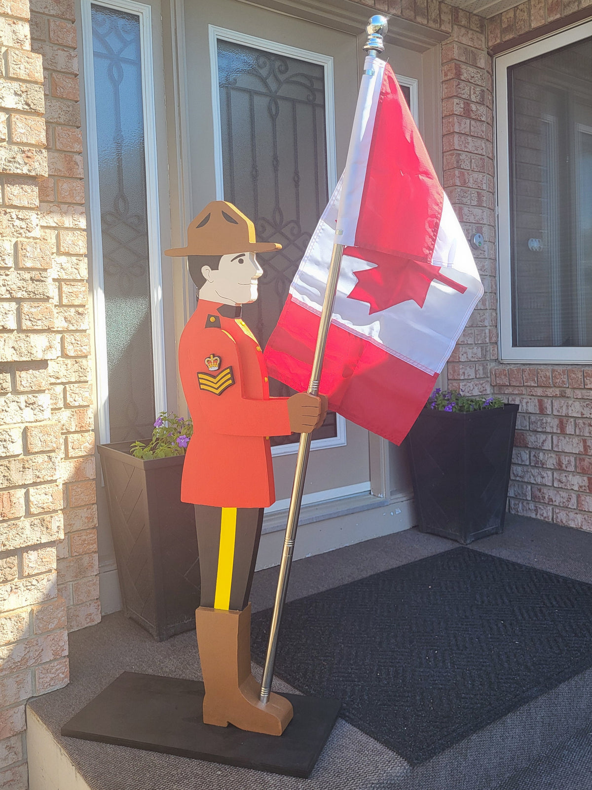 A decorative cutout of a Royal Canadian Mounted Police (RCMP) officer in a red uniform holding a Canadian flag, displayed on a front porch next to a brick wall and door.