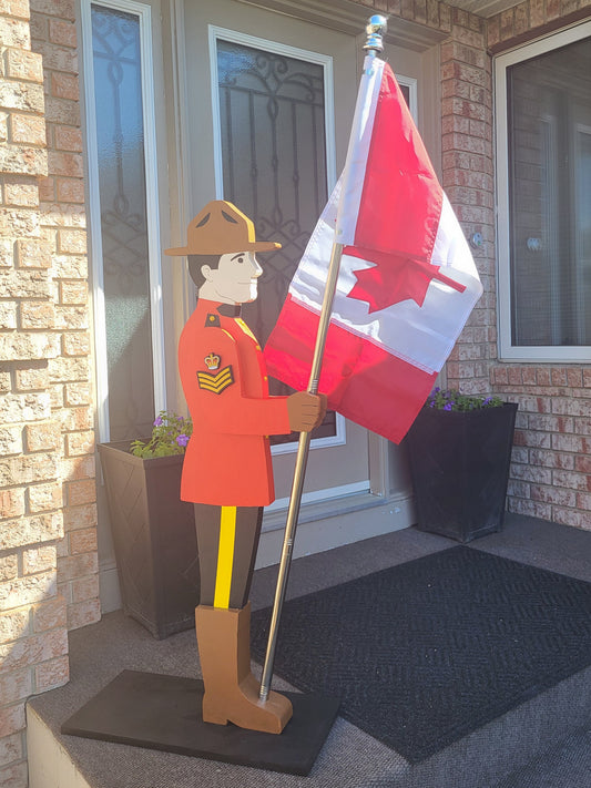 A decorative cutout of a Royal Canadian Mounted Police (RCMP) officer in a red uniform holding a Canadian flag, displayed on a front porch next to a brick wall and door.