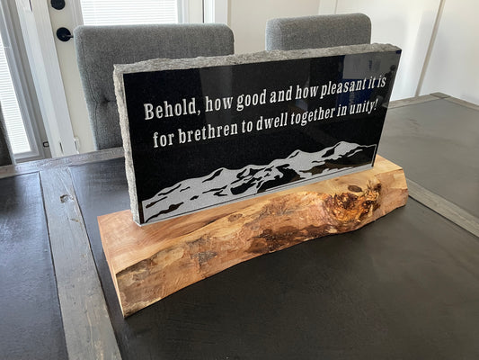 A rectangular black engraved sign with white text reading, 'Behold, how good and how pleasant it is for brethren to dwell together in unity!' above a white mountain silhouette, mounted on a natural live-edge wood base and displayed on a dark table.