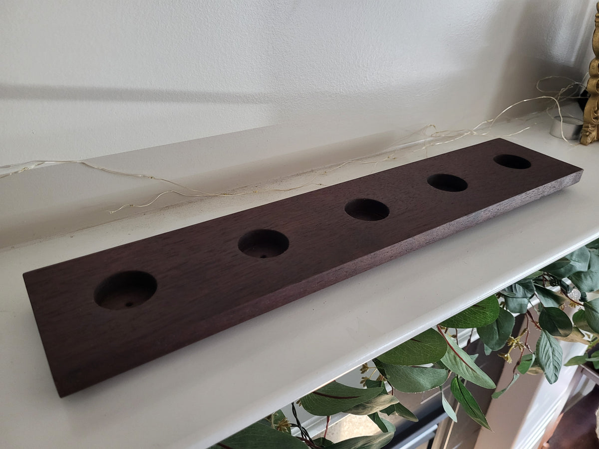 A dark brown wooden rectangular tray with four circular cutouts, resting on a white shelf in an indoor setting with houseplants visible below.